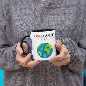 May include: White ceramic mug with a black rim and handle. The mug features a colorful illustration of the Earth and the text "ONE PLANET ONE CHANCE".