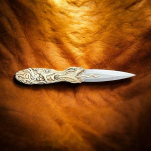 May include: A decorative dagger with a cream-colored handle featuring carved figures and a dragon-like head. The blade is silver and tapers to a sharp point. The dagger is displayed on a brown surface.