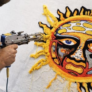 May include: A person using a tufting gun to create a colorful sun design on a white canvas. The sun features a yellow and orange face with blue eyes and black outlines. The tufting gun is silver and blue.