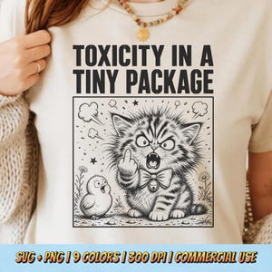 May include: A cream-colored t-shirt featuring a black ink illustration of a grumpy kitten wearing a bow tie and giving the middle finger, standing next to a small chick. The text above reads TOXICITY IN A TINY PACKAGE in bold black letters.