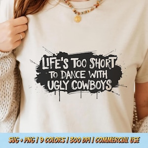 May include: A beige t-shirt with the phrase "LIFE'S TOO SHORT TO DANCE WITH UGLY COWBOYS" in white lettering on a black splatter design. The shirt is a neutral color and has short sleeves. The bottom of the image includes text about the design.