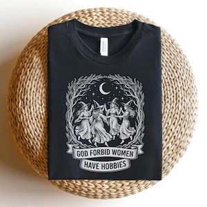 May include: Black t-shirt featuring a white graphic of five witches dancing in a circle under a crescent moon and stars. The text "GOD FORBID WOMEN HAVE HOBBIES" is displayed on a banner below the witches. The shirt is displayed on a woven surface.