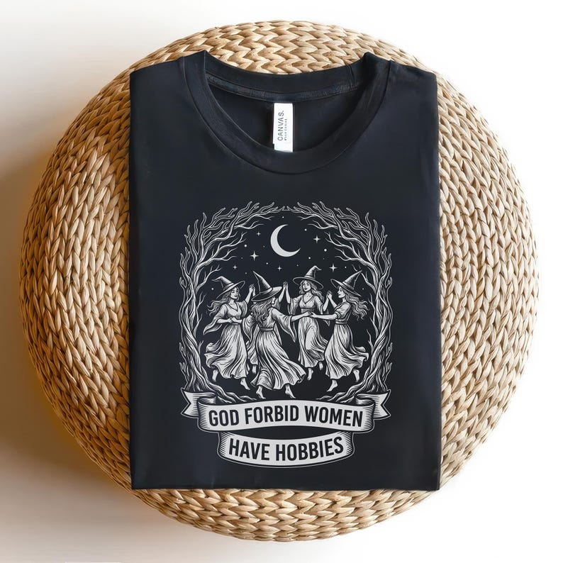 May include: Black t-shirt featuring a white graphic of five witches dancing in a circle under a crescent moon and stars. The text "GOD FORBID WOMEN HAVE HOBBIES" is displayed on a banner below the witches. The shirt is displayed on a woven surface.