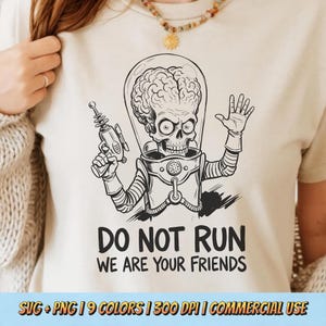 May include: Beige t-shirt featuring a black and white illustration of a Martian with a ray gun, waving. The text "DO NOT RUN WE ARE YOUR FRIENDS" is below the graphic. The shirt is a casual style.