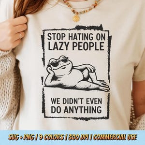 May include: A cream-coloured t-shirt with a black and white graphic of a frog wearing sunglasses, reclining. The text "STOP HATING ON LAZY PEOPLE" and "WE DIDN'T EVEN DO ANYTHING" are printed above and below the frog illustration.