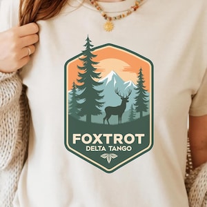 Puede incluir: Una camiseta beige con un diseño gráfico de un ciervo en un entorno de bosque. El gráfico presenta una silueta de un ciervo de pie en un bosque con montañas en el fondo. El texto "FOXTROT DELTA TANGO" está impreso debajo del gráfico.