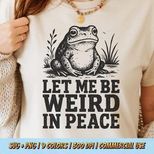 May include: A cream-colored t-shirt featuring a detailed illustration of a frog. The frog is sitting on a patch of grass, with the words "LET ME BE WEIRD IN PEACE" printed below in bold, black letters. The shirt also includes the text "SVG + PNG | 9 COLORS | 300 DPI | COMMERCIAL USE".