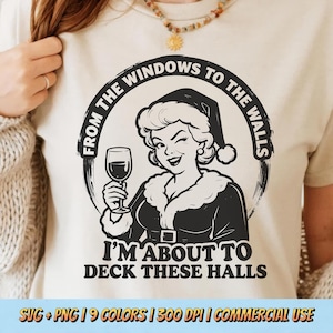 Op de afbeelding: Beige t-shirt met een zwart-witte afbeelding van een vrouw met een kerstmuts die een wijnglas vasthoudt. De tekst rond de afbeelding luidt "FROM THE WINDOWS TO THE WALLS" en "I'M ABOUT TO DECK THESE HALLS".