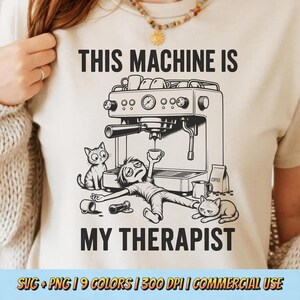 May include: A cream-coloured t-shirt featuring a black graphic of an espresso machine, a tired person lying on the floor, and two cats. The text reads THIS MACHINE IS MY THERAPIST. The design includes a coffee bag, cups, and a portafilter.