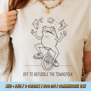 Könnte beinhalten: Beiges T-Shirt mit einer schwarz-weißen Illustration eines Frosches auf einem Einrad, der mit einem Fisch, einer Teetasse und einem Stiefel jongliert. Der Text "OFF TO BEFUDDLE THE TOWNSFOLK" ist darunter gedruckt.