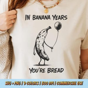 May include: Beige t-shirt with a black and white illustration of a banana holding a balloon. The text "IN BANANA YEARS YOU'RE BREAD" is above and below the banana graphic. The shirt is a neutral color.