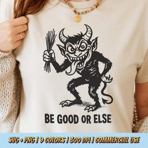 Op de afbeelding: Beige t-shirt met een zwarte illustratie van een duivelachtig wezen met stokken, met de tekst "BE GOOD OR ELSE" eronder. De afbeelding toont een gehoornd wezen met een lange tong. Het t-shirt is neutraal van kleur.