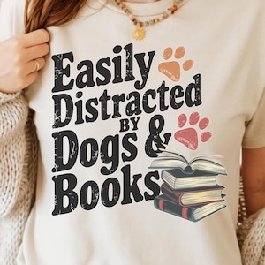 May include: A white t-shirt with the text "Easily Distracted by Dogs & Books" in black. The text is in a vintage style font. There are two paw prints in red and orange on either side of the text. A stack of books is shown below the text.
