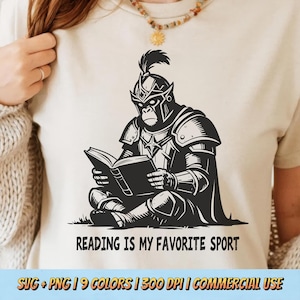 以下が含まれることがあります： クリーム色のTシャツ。黒と白のイラストで、本を読む騎士の鎧を着たゴリラが描かれています。「READING IS MY FAVORITE SPORT」の文字が下にあります。Tシャツはクルーネックです。