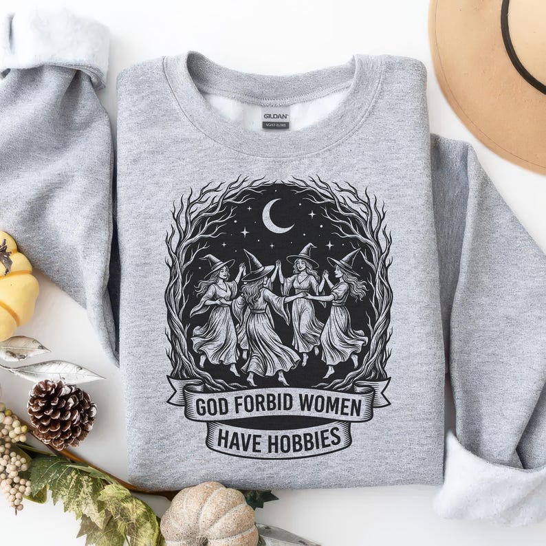 May include: A heather gray sweatshirt featuring a graphic of five witches dancing under a crescent moon, surrounded by a tree branch design. The text "GOD FORBID WOMEN HAVE HOBBIES" is below the graphic.