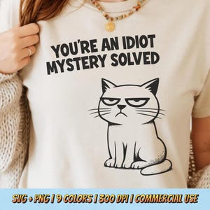 Peut inclure: Un t-shirt beige avec l'inscription noire "YOU'RE AN IDIOT MYSTERY SOLVED". En dessous, un chat de dessin animé noir et blanc avec une expression grincheuse. Le bas de l'image indique "SVG + PNG | 9 COLORS | 300 DPI | COMMERCIAL USE."