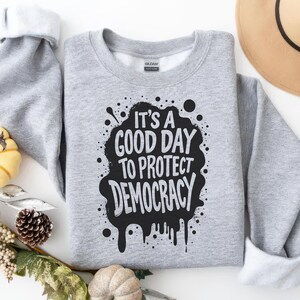 May include: A heather gray sweatshirt with the text "IT'S A GOOD DAY TO PROTECT DEMOCRACY" in a black, distressed design. The sweatshirt has a crew neck and long sleeves. The image includes decorative fall elements.