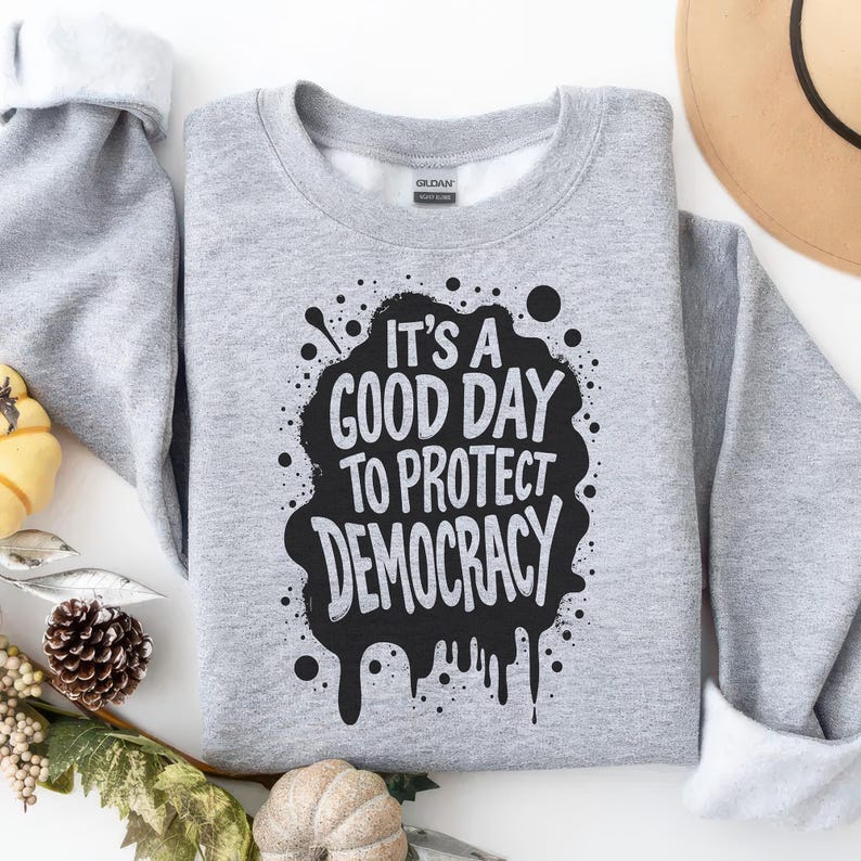 May include: A heather gray sweatshirt with the text "IT'S A GOOD DAY TO PROTECT DEMOCRACY" in a black, distressed design. The sweatshirt has a crew neck and long sleeves. The image includes decorative fall elements.