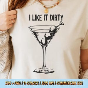 Peut inclure: T-shirt beige avec l'inscription "I LIKE IT DIRTY" au-dessus d'une illustration en noir et blanc d'un verre à martini avec des olives. Le t-shirt a un col rond et des manches courtes. L'image comprend également le texte "SVG + PNG | 9 COLORS | 300 DPI | COMMERCIAL USE".