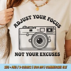 May include: Beige t-shirt with a black graphic of a vintage camera and the text "ADJUST YOUR FOCUS NOT YOUR EXCUSES". The shirt is a neutral color and features a classic camera design.