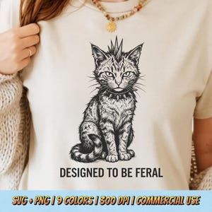 May include: A cream-colored t-shirt featuring a detailed black and white illustration of a cat with a mohawk and studded collar. The text "DESIGNED TO BE FERAL" is printed below the cat image. The shirt is a casual wear item.