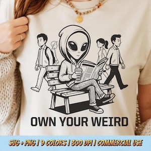 Puede incluir: Camiseta beige con un gráfico en blanco y negro de un extraterrestre leyendo un periódico sentado en un banco. El texto "OWN YOUR WEIRD" está debajo del gráfico. Otras personas caminan en el fondo.