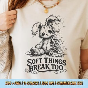 May include: A cream-coloured t-shirt with a black and white illustration of a stitched bunny, one side disintegrating. The text "SOFT THINGS BREAK TOO" is below the bunny. The bottom of the shirt has text that reads "SVG + PNG | 9 COLORS | 300 DPI | COMMERCIAL USE".