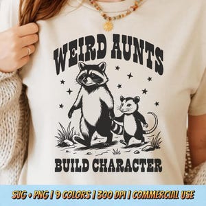 以下が含まれることがあります： ベージュ色のTシャツ。黒と白のイラストでアライグマとオポッサムが描かれ、「WEIRD AUNTS BUILD CHARACTER」の文字。デザインには星と草が含まれています。シャツは明るい色です。