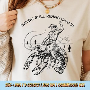 Peut inclure: T-shirt crème avec une illustration en noir et blanc d'un cow-boy chevauchant une écrevisse géante. Le texte "BAYOU BULL RIDING CHAMP" est au-dessus de l'image. Le t-shirt comprend également le texte "SVG + PNG | 9 COLORS | 300 DPI | COMMERCIAL USE".