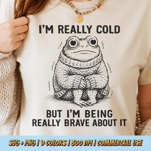 Op de afbeelding: Beige T-shirt met een zwarte illustratie van een kikker met een trui. De tekst op het shirt luidt "I'M REALLY COLD BUT I'M BEING REALLY BRAVE ABOUT IT". De onderkant van het shirt heeft de tekst "SVG + PNG | 9 COLORS | 300 DPI | COMMERCIAL USE".