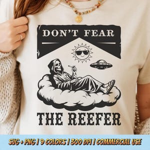 May include: Cream-coloured t-shirt with a graphic of the Grim Reaper reclining on a cloud, smoking. The text above reads "DON'T FEAR" and below, "THE REEFER." A sun wearing sunglasses and a UFO are also depicted. The shirt is a light colour.