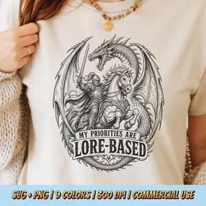 以下が含まれることがあります： ベージュ色のTシャツ。白黒のイラストで、馬に乗った騎士、ドラゴン、そして「MY PRIORITIES ARE LORE-BASED」の文字が描かれています。Tシャツの下部には「SVG + PNG | 9 COLORS | 300 DPI | COMMERCIAL USE」の文字があります。