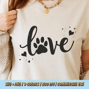 May include: Cream-colored t-shirt with the word "love" in a black, cursive font, featuring a paw print in place of the "o". Small black hearts and speckles surround the word. The bottom of the shirt has the text "SVG + PNG | 9 COLORS | 300 DPI | COMMERCIAL USE".