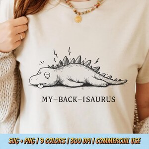 Może przedstawiać: Beżowa koszulka z czarno-białą ilustracją zmęczonego dinozaura i napisem "MY-BACK-ISAURUS". Koszulka ma okrągły dekolt i krótkie rękawy. Na dole koszulki znajduje się napis "SVG + PNG | 9 COLORS | 300 DPI | COMMERCIAL USE".