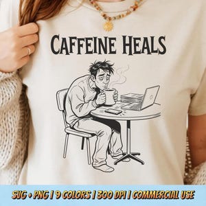 Puede incluir: Camiseta beige con la frase "Caffeine Heals" sobre una ilustración en blanco y negro de una persona cansada bebiendo café. La imagen muestra a una persona en una mesa con un portátil y un teléfono.