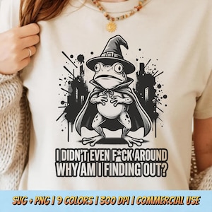 May include: Beige t-shirt featuring a cartoon frog dressed as a wizard, wearing a hat and cape. The frog is set against a black cityscape and the text reads, "I DIDN'T EVEN F*CK AROUND WHY AM I FINDING OUT?"