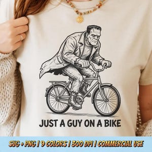 May include: A cream-colored t-shirt featuring a black line-art illustration of a creature with stitches riding a bicycle. Below the graphic, the text reads JUST A GUY ON A BIKE. The design is centered on the chest of the garment.
