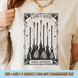 May include: Cream-colored t-shirt featuring a black and white graphic of six witch brooms. The text "A Girl's Gotta Have Options" is displayed above and below the brooms in a decorative frame. The shirt is a light neutral color.