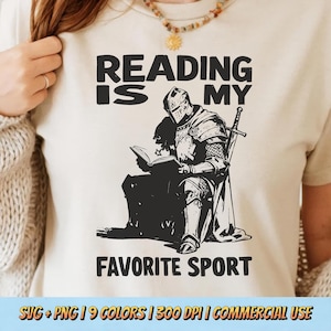 May include: A cream-coloured t-shirt featuring a black graphic that reads "READING IS MY FAVORITE SPORT." The graphic shows a knight reading a book, with a sword beside him. The shirt is being worn by a person.