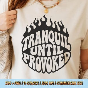Puede incluir: Camiseta color crema con un diseño gráfico negro. El diseño es una forma de llama circular con las palabras "TRANQUIL UNTIL PROVOKED" en una fuente retro audaz. Es una prenda informal.