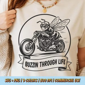 Puede incluir: Camiseta beige con un gráfico en blanco y negro de una abeja montando una motocicleta. La abeja lleva gafas y el texto "BUZZIN THROUGH LIFE" está en una pancarta debajo. La camiseta tiene cuello redondo.