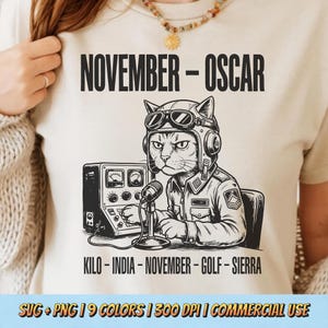 以下が含まれることがあります： ベージュ色のTシャツ。黒と白のイラストで、パイロットのヘルメットとジャケットを着た猫がラジオを操作しています。「NOVEMBER - OSCAR」の文字が猫の上に、「KILO - INDIA - NOVEMBER - GOLF - SIERRA」の文字が下にあります。Tシャツには「SVG + PNG | 9 COLORS | 300 DPI | COMMERCIAL USE」の文字も入っています。