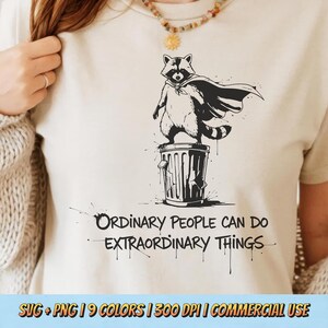 May include: Cream-coloured t-shirt with a black and white illustration of a raccoon in a cape on a bin. The text "ORDINARY PEOPLE CAN DO EXTRAORDINARY THINGS" is printed below the image.