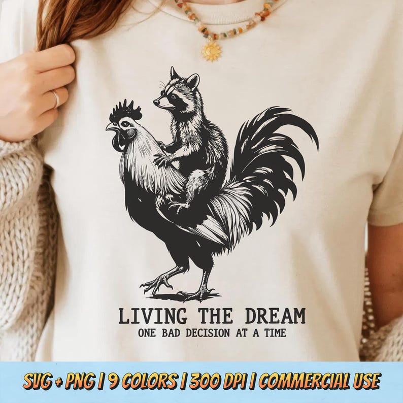 May include: Cream-colored t-shirt featuring a black and white illustration of a raccoon riding a rooster. The text "LIVING THE DREAM" and "ONE BAD DECISION AT A TIME" are printed below the image. The shirt is a graphic tee.