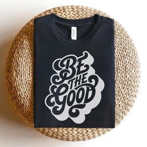 May include: Black t-shirt with the phrase "BE THE GOOD" in a white, stylized font. The shirt is displayed on a round, woven wicker surface. The shirt has a crew neck and a tag.