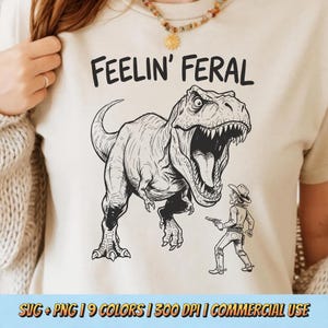 May include: Beige t-shirt with a black graphic of a Tyrannosaurus Rex facing a cowboy with a gun. The text "FEELIN' FERAL" is above the dinosaur. The shirt is a light color.