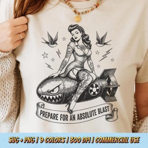 May include: A cream-colored t-shirt featuring a black ink graphic of a pin-up figure sitting on a cartoon bomb with a shark-like face. The design includes swallows, stars, lightning bolts, and a banner with the text PREPARE FOR AN ABSOLUTE BLAST.