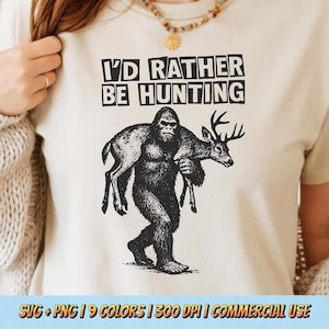 May include: Beige t-shirt with the text "I'D RATHER BE HUNTING" in black block letters. A black and white graphic depicts a Bigfoot carrying a deer. The shirt is a casual wear item.
