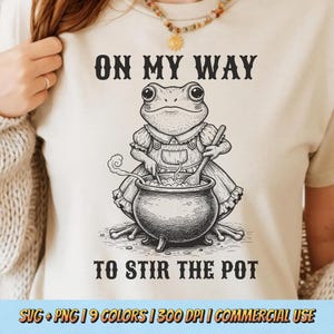 May include: Beige t-shirt featuring a whimsical illustration of a frog stirring a cauldron. The text "ON MY WAY TO STIR THE POT" is above and below the frog. The design is in black and white.