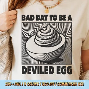 May include: A cream-colored t-shirt with a black and white graphic of a deviled egg. The text "BAD DAY TO BE A DEVILED EGG" is above and below the graphic. The shirt is being worn by a person.